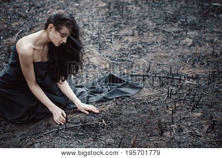 Sad girl sitting on the burned-out land. Concept of environmental disaster. Global problems with ecology. Young attractive multi-racial Asian Caucasian woman in black dress on nature