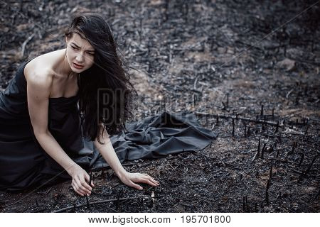Sad girl sitting on the burned-out land. Concept of environmental disaster. Global problems with ecology. Young attractive multi-racial Asian Caucasian woman in black dress on nature