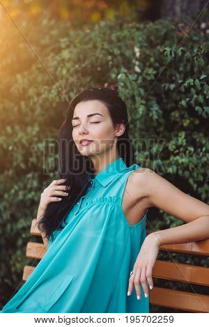 Beautiful young woman with long hair in dress sitting on a bench in a city park. Attractive young multi-racial Asian Caucasian gorgeous girl model resting with closed eyes outdoors