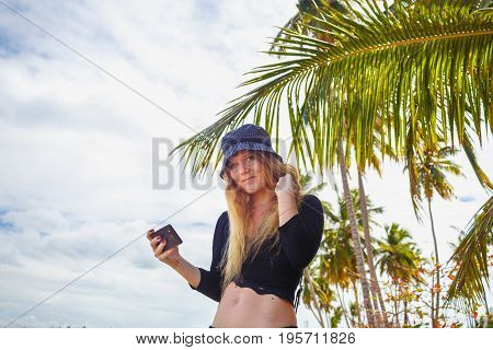 Pretty girl the girl in the hat holding smartphone under palm leaves