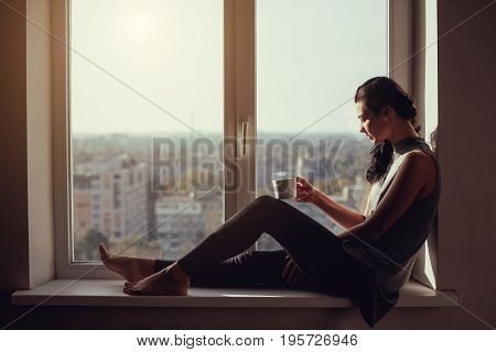 Resting and thinking woman. Calm girl with cup of tea or coffee sitting on the window-sill at home. Side view. Young attractive multi-racial Asian Caucasian lady in casual clothes resting indoors