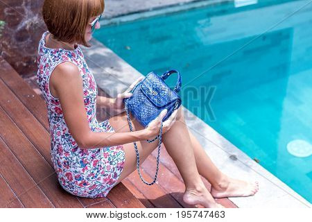 Summer portrait of a beautiful sexy girl with sunglasses and luxury handmade snakeskin python handbag in the swimming pool. Fashion outdoor. Tropical Bali island.