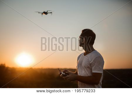Silhouette of a man piloting drone in the air with a remote controller in his hands on sunset. Pilot takes aerial photos and videos with quadcopter from above. Copter on background