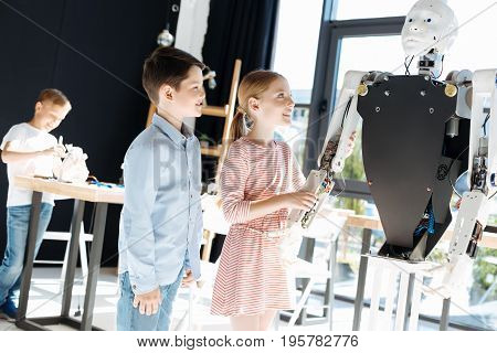 Awe-inspiring robot. Fascinated lovely children standing near a big human robot and checking out its work while the girl holding its hand