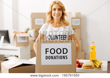 You can add something. Admirable young devoted lady holding a box full of food while working pro bono and packing parcels for shipment
