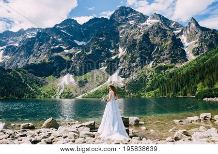 the bride in a beautiful blue dress and a wreath of white flowers holding a bouquet of purple flowers and eucalyptus overlooking the green mountains and lake