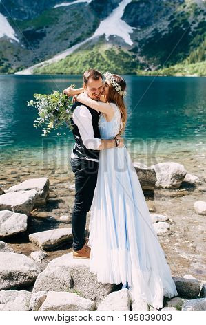 bride with the beautiful blue dress and groom hugging with views of the beautiful green mountains and lake with blue water