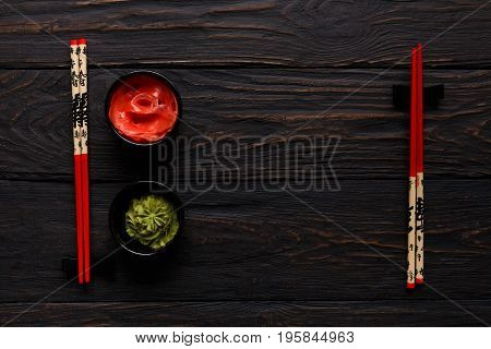 Pieces of traditional japanese tablewear. Red chopsticks with Bon appetite written in hierogliphs, small bowls with ginger and wasabi on dark wooden table. Top view, flat lay, copy space, background