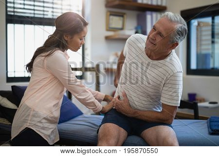 Senior male patient sitting on bed while female therapist examining back at hospital ward