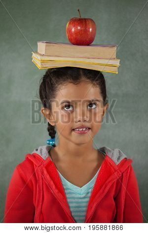 Young girl balancing books and apple on her head against chalk board