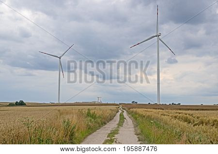 Two wind turbines electric. In the middle of an unpaved, sandy road. Between the ruts and the sides grow green grass. In the distance to the left industrial constructions. On both sides crop fields on which grow ripe yellow corn. The road is winding. It's
