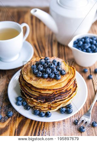 Pancakes healthy breakfast with blueberries, bog whortleberry, cup of green tea, cup of blueberries and teapot on brown wooden background