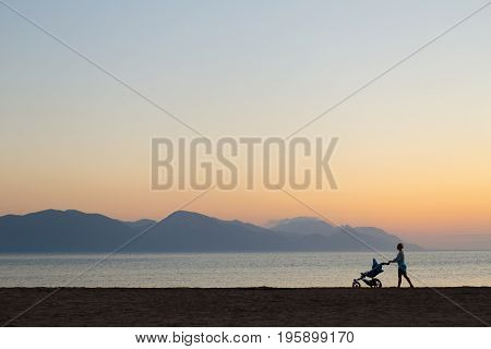 Silhouette of mother with stroller enjoying motherhood at sunset landscape. Walking or jogging woman with pram on a beach at sunset. Beautiful inspirational mountains landscape.
