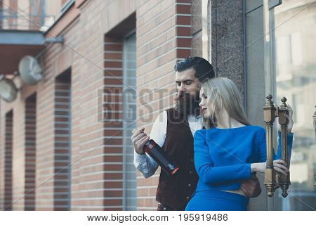 Girlfriend and boyfriend looking at bottle of wine. Pretty woman or girl in blue dress and man or hipster outdoors on brick wall. Alcohol and convive. Couple in love. Unhealthy lifestyle. Bad habits