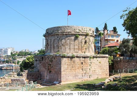 Antalya, Turkey - 1 Jun 2017: The rocky coast of city with the ancient roman Hidirlik Tower with turkish red flag, historically used as the fortification and the lighthouse on a blue sky background.