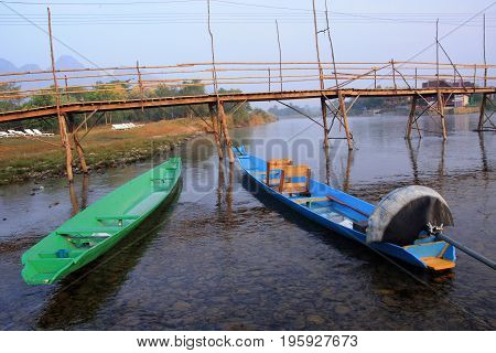 a blue boat and a green boat float side by side in a small river under a wooden bridge.