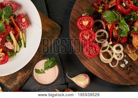 Close-up of a fresh salad with chicken breast bell pepper onion garlic and tomato on round wooden board. Diet rye bread and fresh sauce on dark stone background. Top view