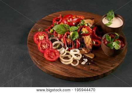 Braised chicken breast with baked vegetables and сhili sauce in a bowl on round wooden board over dark stone background. close-up. horizontal. Healthy low-calories food.