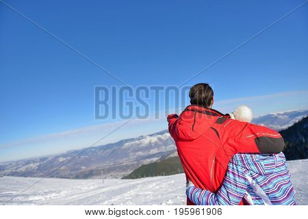 Rear view of a loving couple in fur hood jackets looking at snowed mountain range.