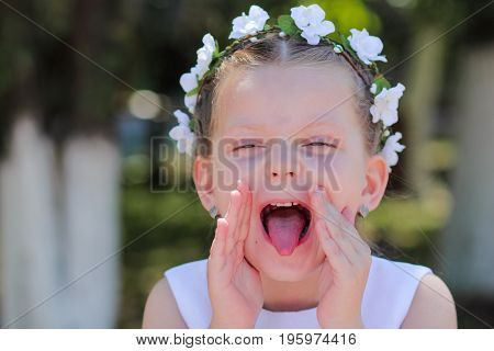 little girl showing tongue and covers her face with his hands a child with a wreath of artificial flowers on her head