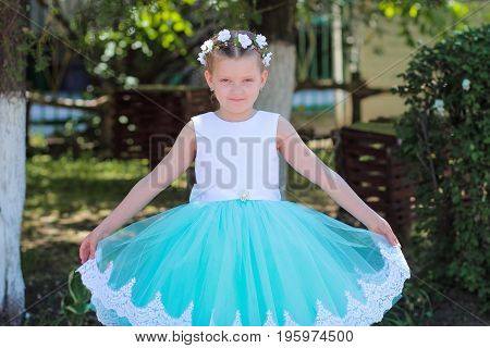 Small cute girl in a blue and white dress and wreath raises the hem of her dress