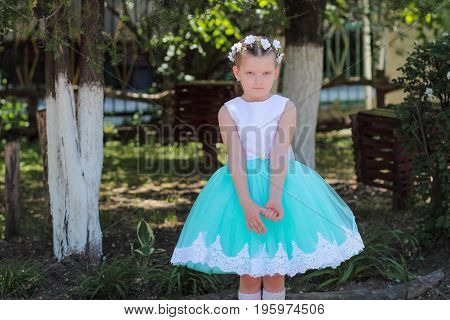 Offended child small cute girl in a blue and white dress child with a wreath of artificial flowers on her head