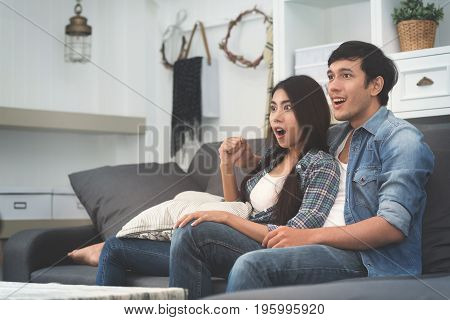 Cheerful beautiful young Asian couple watching TV at home.