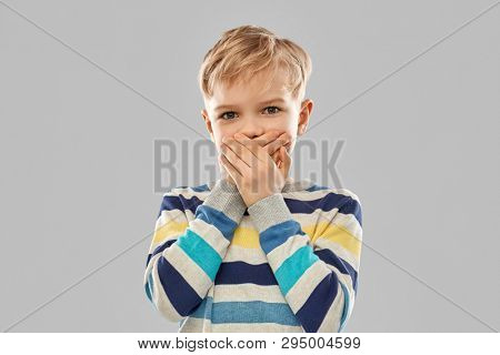 childhood, expressions and people concept - shocked little boy in striped pullover closing his mouth by hands over grey background