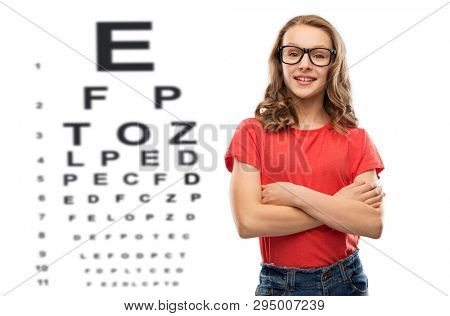 vision, eyesight and eyewear concept - smiling teenage student girl in glasses and red t-shirt with crossed arms over eye test chart background