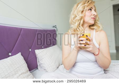 Attractive Blonde Woman Sits On The Edge Of The Bed At Home And Holds A Stocker With Orange Juice In