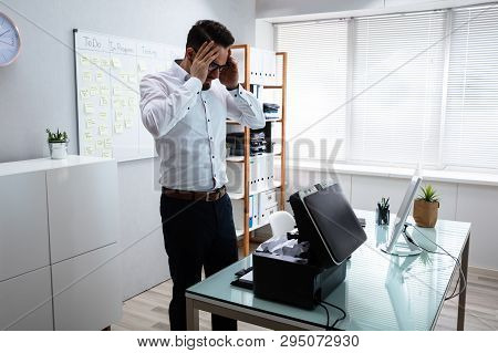 Young Irritated Businessman Looking At Printer In Office