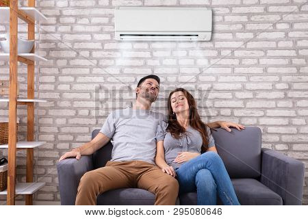 Happy Young Couple Sitting Under Air Conditioner On Couch At Home