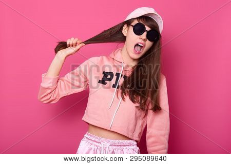 Portrait Of Charming Dark Haired Female In Pink Hoodie, Cap And Suglasses, Holding Pulling Hair, Loo