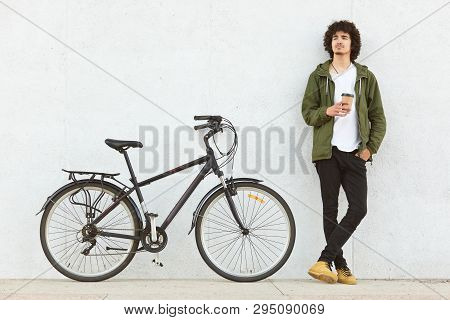 Indoor Shot Of Attractive Pencive Man With Crisp Hair Drinking Hot Baverage From Paper Cup, Looks Th