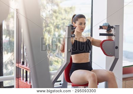 Photo Of Beautiful Young Girl Performing Weight Lifting To Improve Her Upper Body On Machine In Gym.