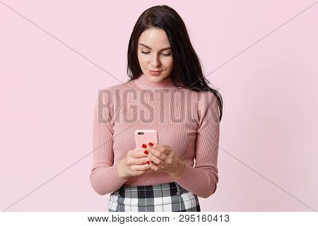 Sweet Attractive Smiling Young Lady Stands Isolated Over Pink Background, Looking At Her Mobile Phon