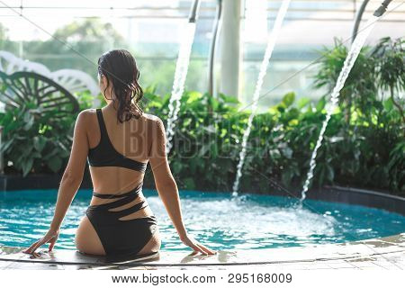 Shoot From Back Of Sexy Slim Female In Swimsuit Sits On Edge Of Swimming Pool Between Green Bushes O