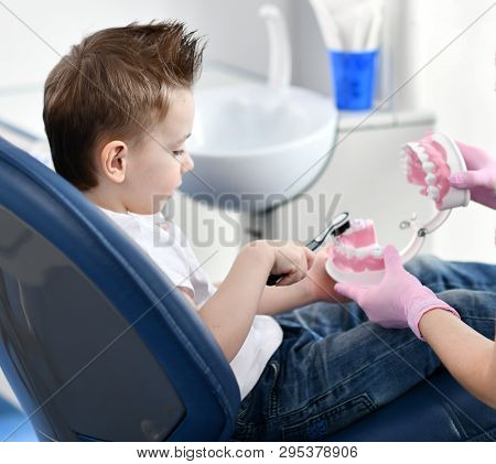 Kid Boy In Dental Chair Trains To Clean Teeth With A Dummy Mandibula Jaw On Background With Dental O