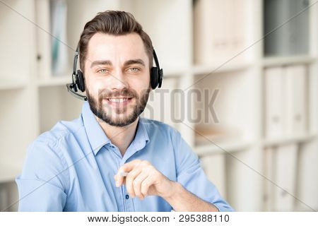 Smiling young operator with headphone looking at you while working with clients questions and inquiries in office