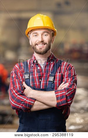 Happy young smiling engineer or machinebuilder in hardhat and overalls crossing his arms on chest