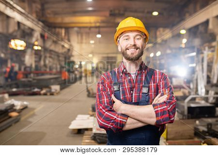 Young smiling professional in overalls and protective helmet standing in front of camera inside large machinebuilding plant