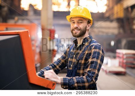 Happy young successful factory engineer in protective helmet looking at you while controling working process