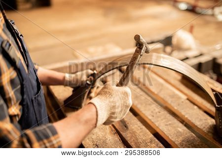 Gloved engineer or machinebuilder using hammer while holding metallic workpiece on wooden workbench