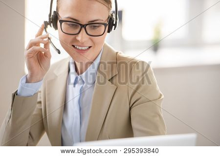 Content confident female call center operator in glasses looking at computer screen while answering call using hands-free device in office