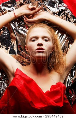 Arty Portrait Of A Fashionable Red-haired Model In Red With Hands Up Over Wrinkled Silver Foil Backg