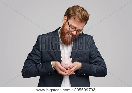 Handsome Businessman With Ginger Beard Smiling And Rocking Small Moneybox With Bank Investments Agai