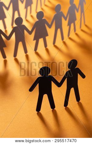 Black paper figure of male couple in front of a crowd of paper people holding hands on orange surface. Social movement, protest, minorities concept.