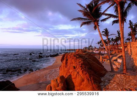 Cabo San Lucas Resort Shore At Sunrise With Rocks, Ocean, Palm Trees And Sand