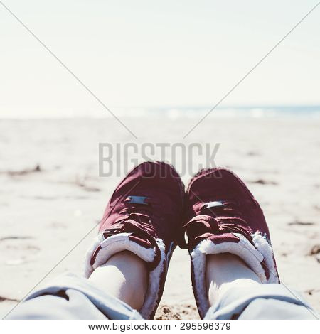 Feet Of Young Woman In Sport Footwear And Jeans Relaxing After Hiking On Sand Beach. Square Pov. Adv