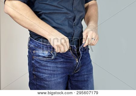 Man Unzipps Blue Jeans. Man Torso, Fingerring, Blue Shirt, Casual Wear. Indoors, Grey Empty Backgrou
